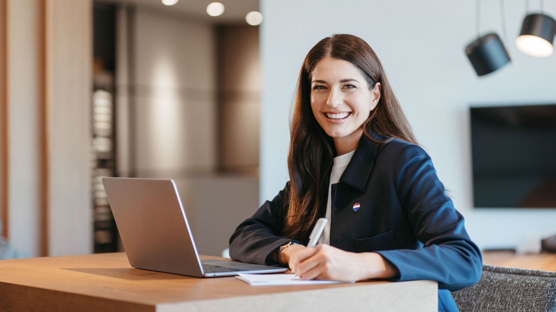 Agente immobiliare donna sorridente al computer con giacca e spilla REMAX che guarda in camera
